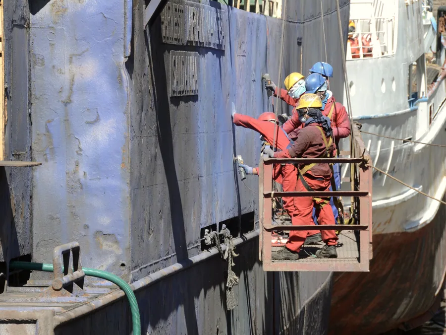 Crew in coveralls and hard hats coating a vessel hull from a cradle